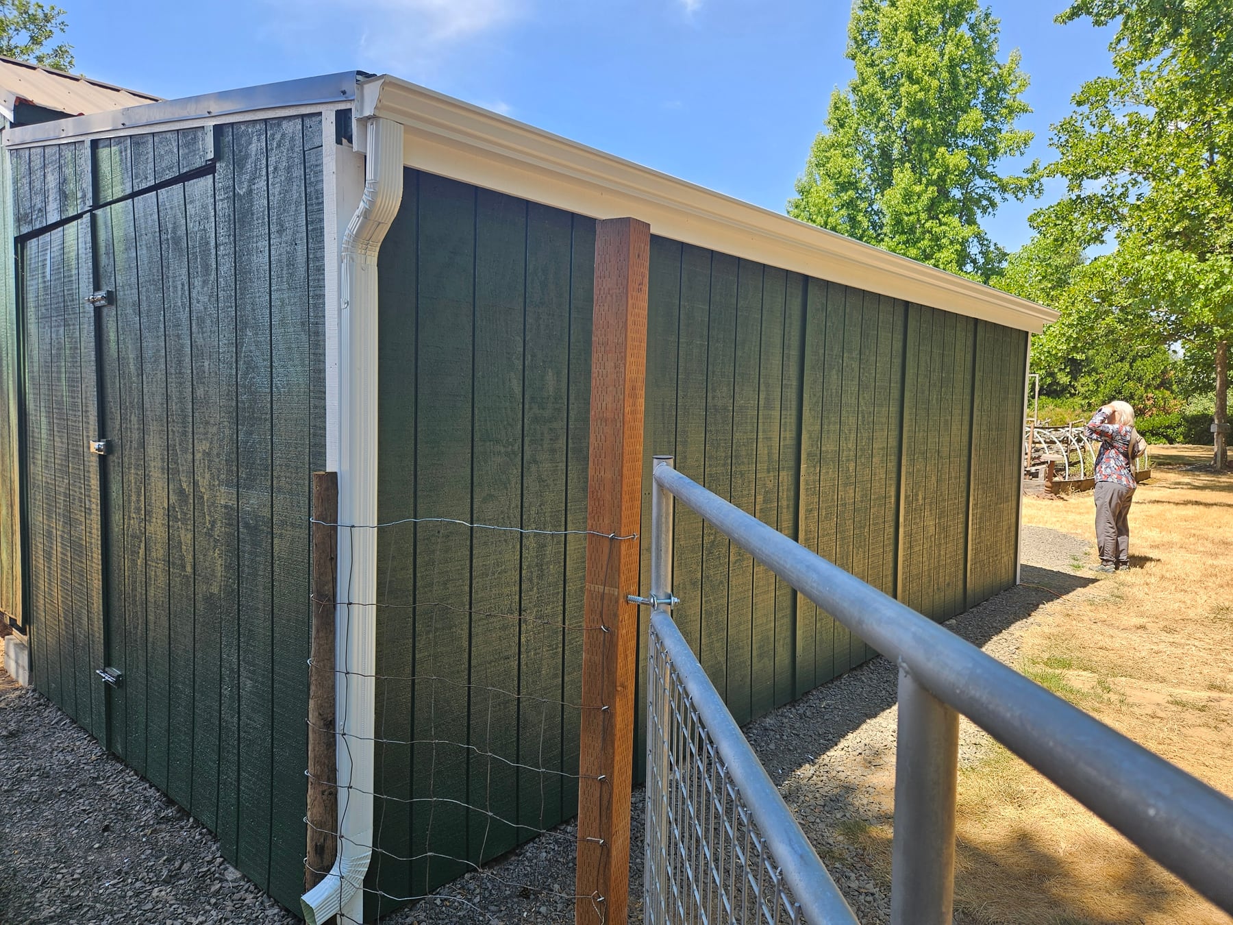White gutter and downspout installed on a green outbuilding beside a fenced area